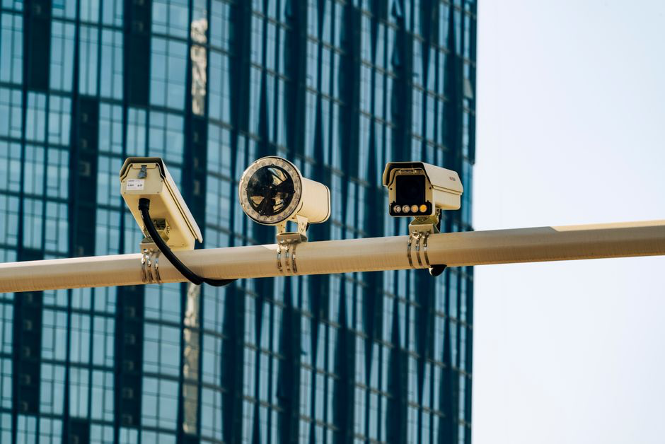 Three surveillance cameras mounted on a beam against a glass office tower